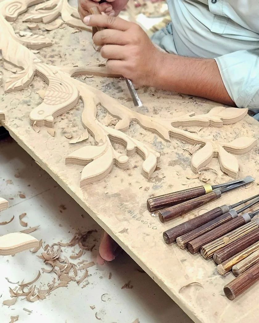 Tablero con ornamento vegetal tallado en relieve, virutas de madera y una hilera de gubias de distintos anchos apoyadas al lado